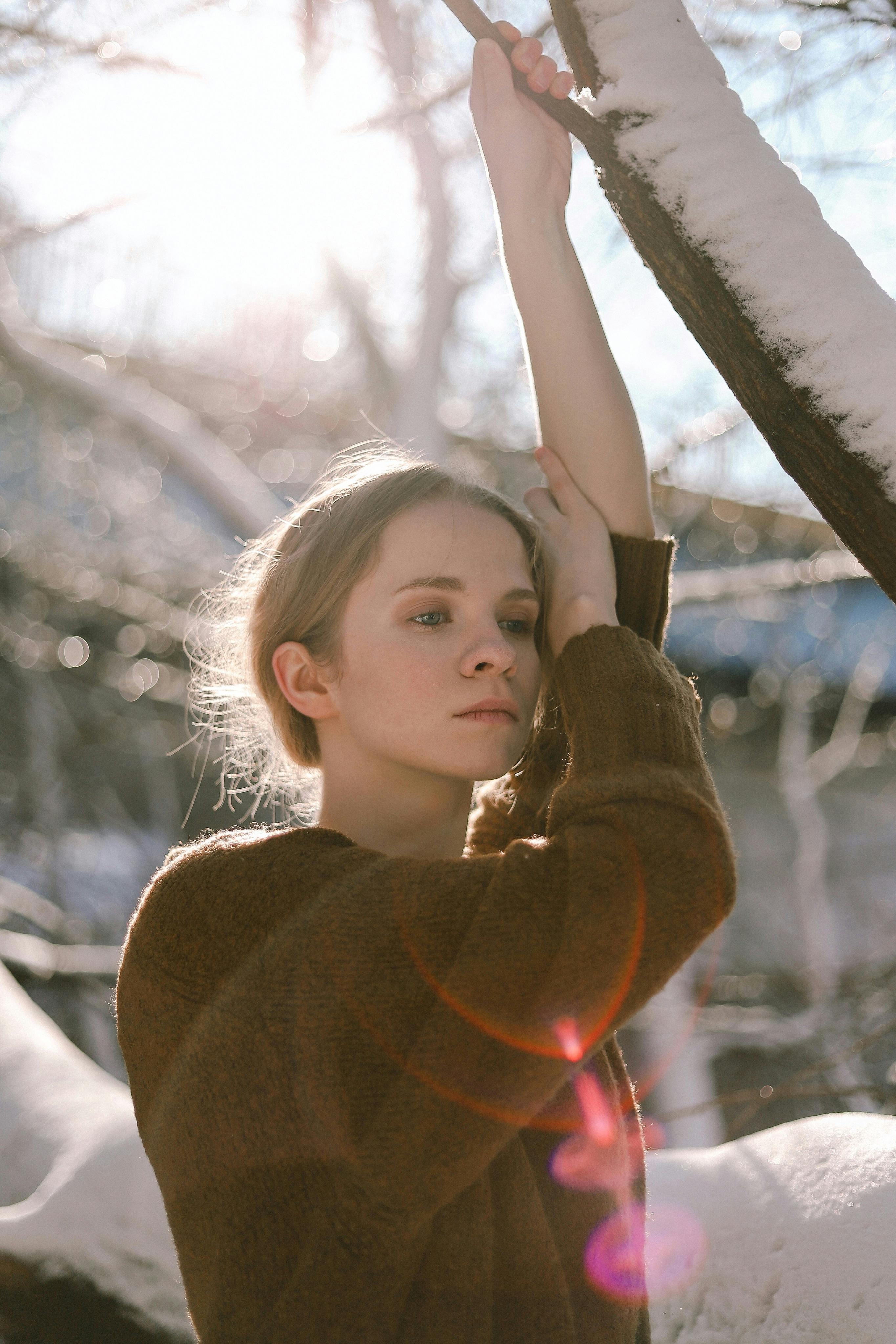 A female therapy client for anxiety and perfectionism stands alone in the winter, looking
                somber. She is wearing a red sweater and is touching a tree.