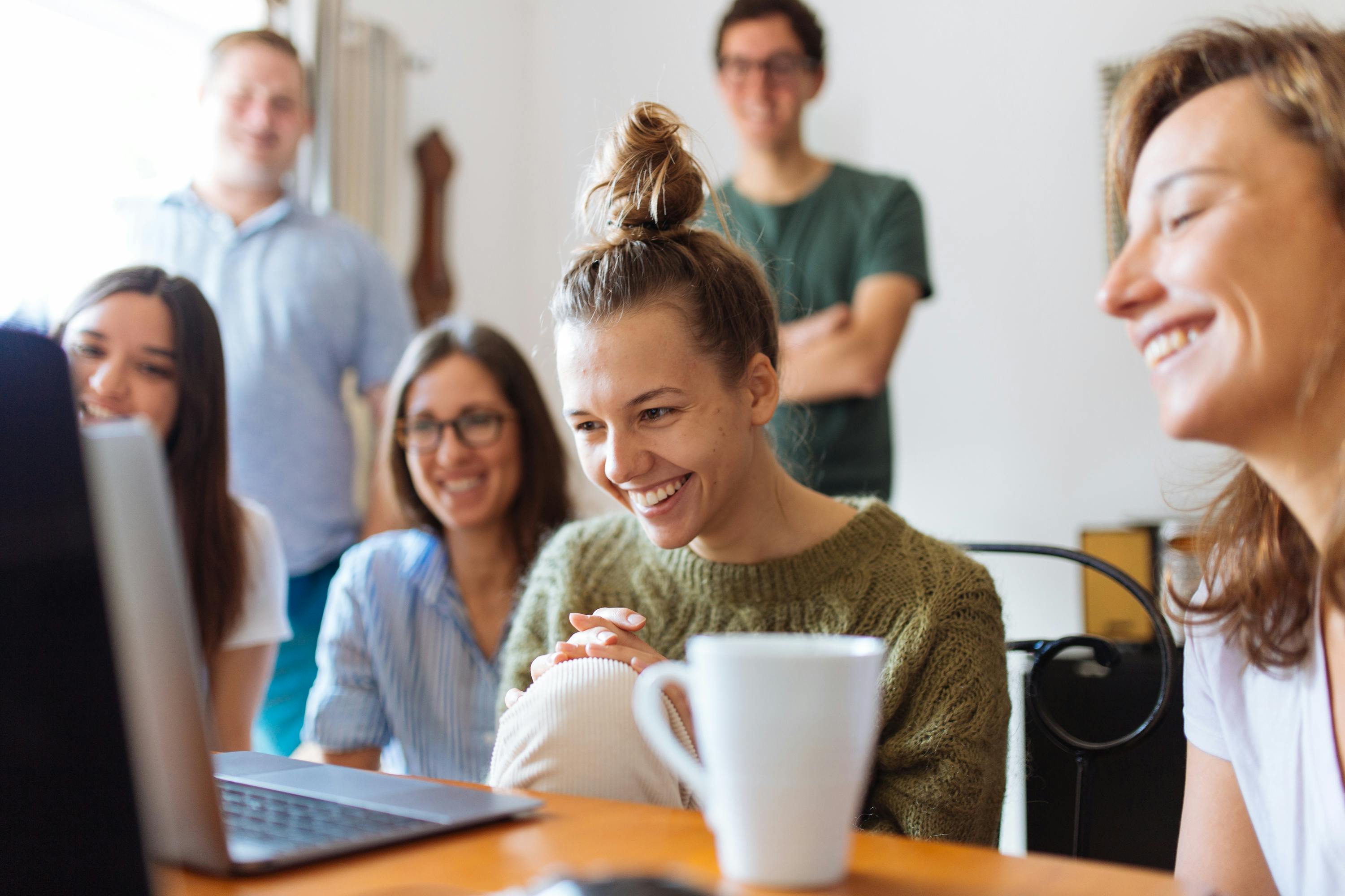 A young adult woman is in the workplace surrounded by co-workers. She is smiling and happy.