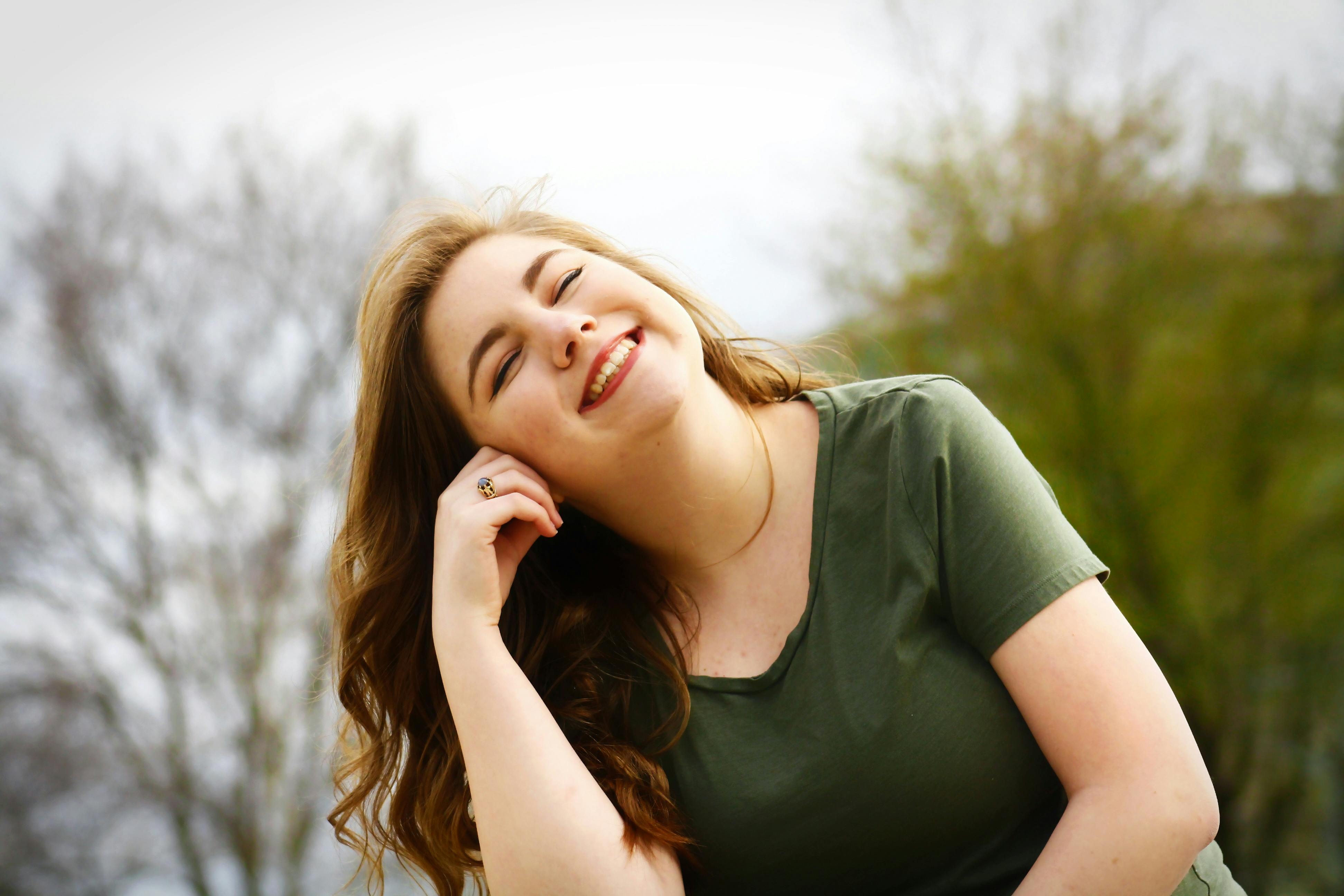 A young woman is seated and smiling outdoors. She is wearing a green shirt and appears happy and at ease.