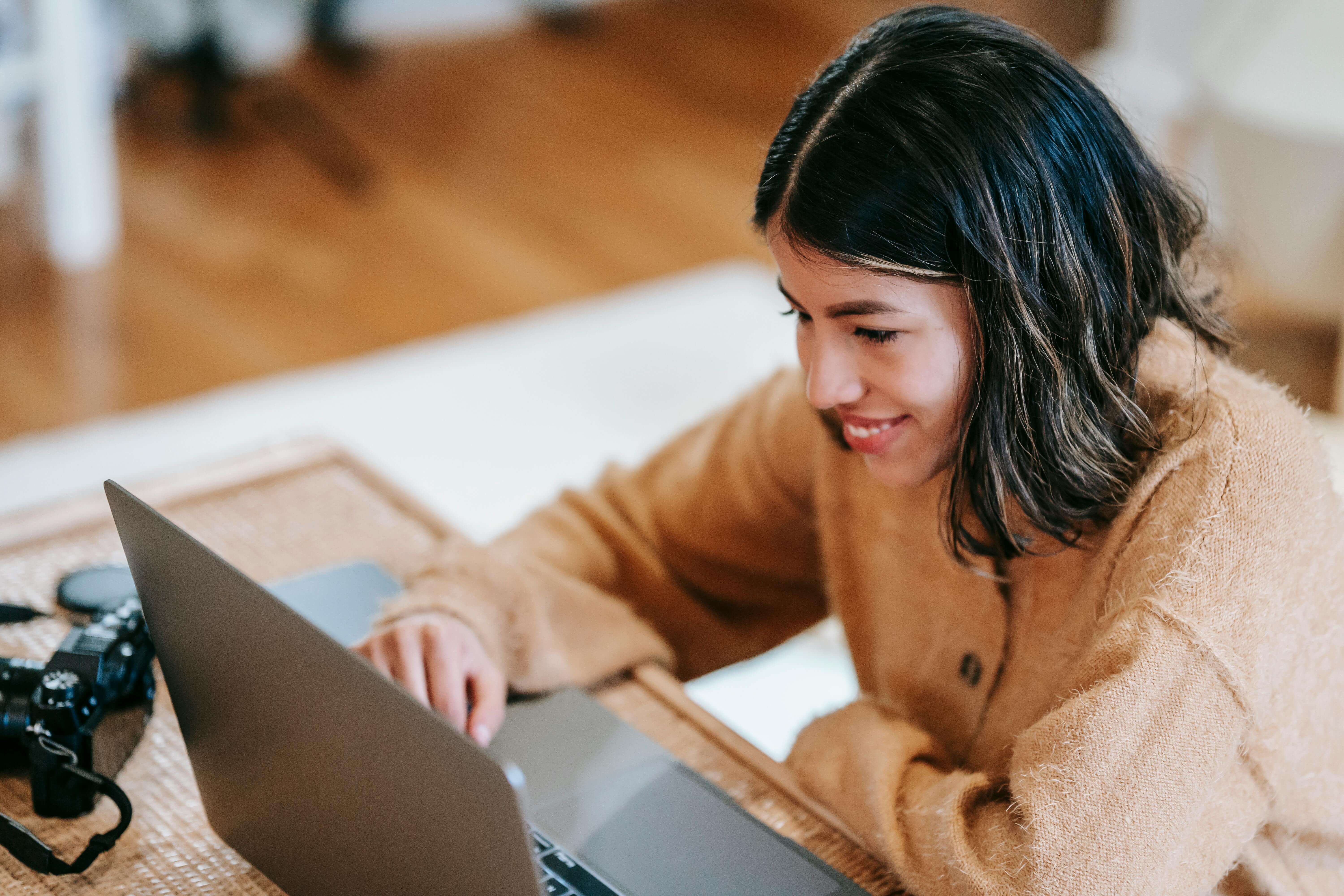 A brunette girl wearing a camel sweater is smiling at her laptop. She appears engaged
                and happy following a perfectionism therapy telehealth appointment.