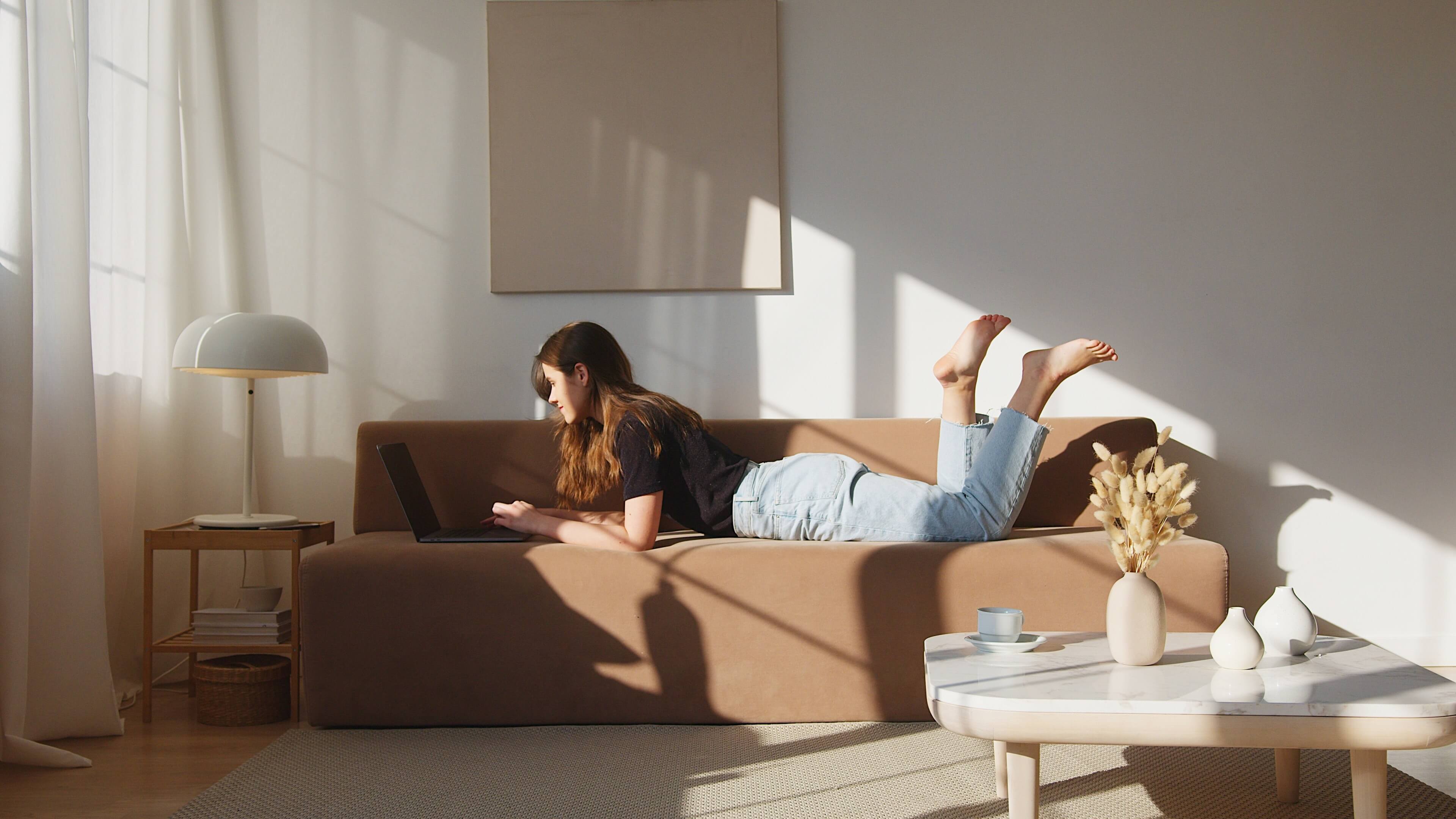 A woman is seated on her couch at home, using her computer. She is comfortable and relaxed.